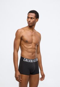 Μαύρο βαμβακερό μποξεράκι με λευκό λογότυπο "CALVIN KLEIN" στη μέση, εφαρμοστή σχεδίαση και λεία υφή. Το μοντέλο στέκεται με γυμνό κορμό.