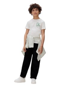 Camiseta blanca con logo verde, camisa a rayas verde y blanca atada a la cintura, pantalones negros y zapatillas blancas. Cabello rizado.