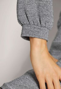 Grau strukturierter Ärmel eines Kleidungsstücks mit gerafftem Bund. Die sichtbare Hand zeigt glatte Haut. Der Stoff weist ein Strickmuster auf.