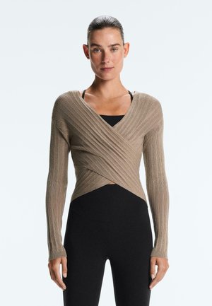 Hátul összekötött hajjal rendelkező nő, bézs színű bordázott kötött pulóverrel, amely fekete felső fölött van, és magas derekú fekete leggingset visel, fehér háttér előtt.