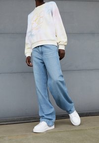 Pastelkleurige tie-dye sweatshirt met een bloemenafbeelding, gecombineerd met lichtblauwe wijde jeans en witte sneakers. Gestructureerde, casual outfit.