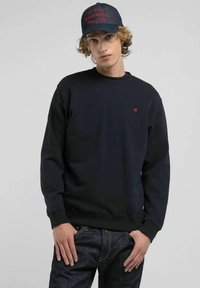 Felpa blu navy con girocollo, polsini e orlo a coste; presenta un piccolo logo rosso sul petto. Abbinata a jeans in denim scuro e un cappellino.