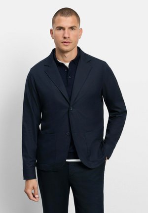 Mann trägt einen eng anliegenden dunkelblauen Blazer über einem dunkelblauen Poloshirt und schwarzen Hosen, steht vor einem schlichten weißen Hintergrund.