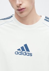 Biele atletické tričko s modrým logom Adidas, s modrými pruhmi na pleciach a okrúhlym výstrihom, vyrobené z hladkej, ľahkej látky.