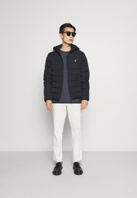 Doudoune marine avec une fermeture éclair, des poches avant et une capuche. Portée par-dessus une chemise bleue texturée et un pantalon blanc, complétée par des chaussures noires.