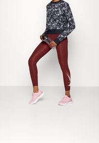 Schwarz- und grau gemustertes Langarmoberteil, maroonfarbene Leggings mit weißen Akzenten und pinkfarbene Sportschuhe. Das Model hält eine durchsichtige Wasserflasche.