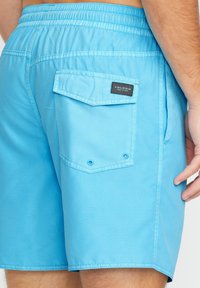 Shorts de bain bleu clair avec taille élastique, poche latérale et poche arrière avec une étiquette noire. Texture lisse et coupe ajustée.