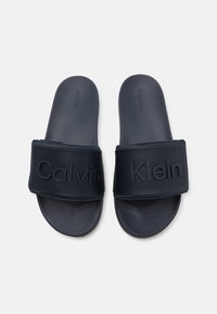 Fekete csúszásmentes papucsok texturált pánttal, amelyen a "Calvin Klein" felirat van belefúrva. Mesterséges anyagból készült, sima talpbéléssel.