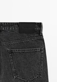 Close-up of black denim jeans showing back pocket and black leather brand tag on waistband, "Massimo Dutti" embossed.