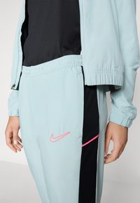 Compleu de trening bleu deschis cu talie elastică și panou negru; are logo-ul roz Nike și o fâșie subțire roz pe lateral. Textură netedă.