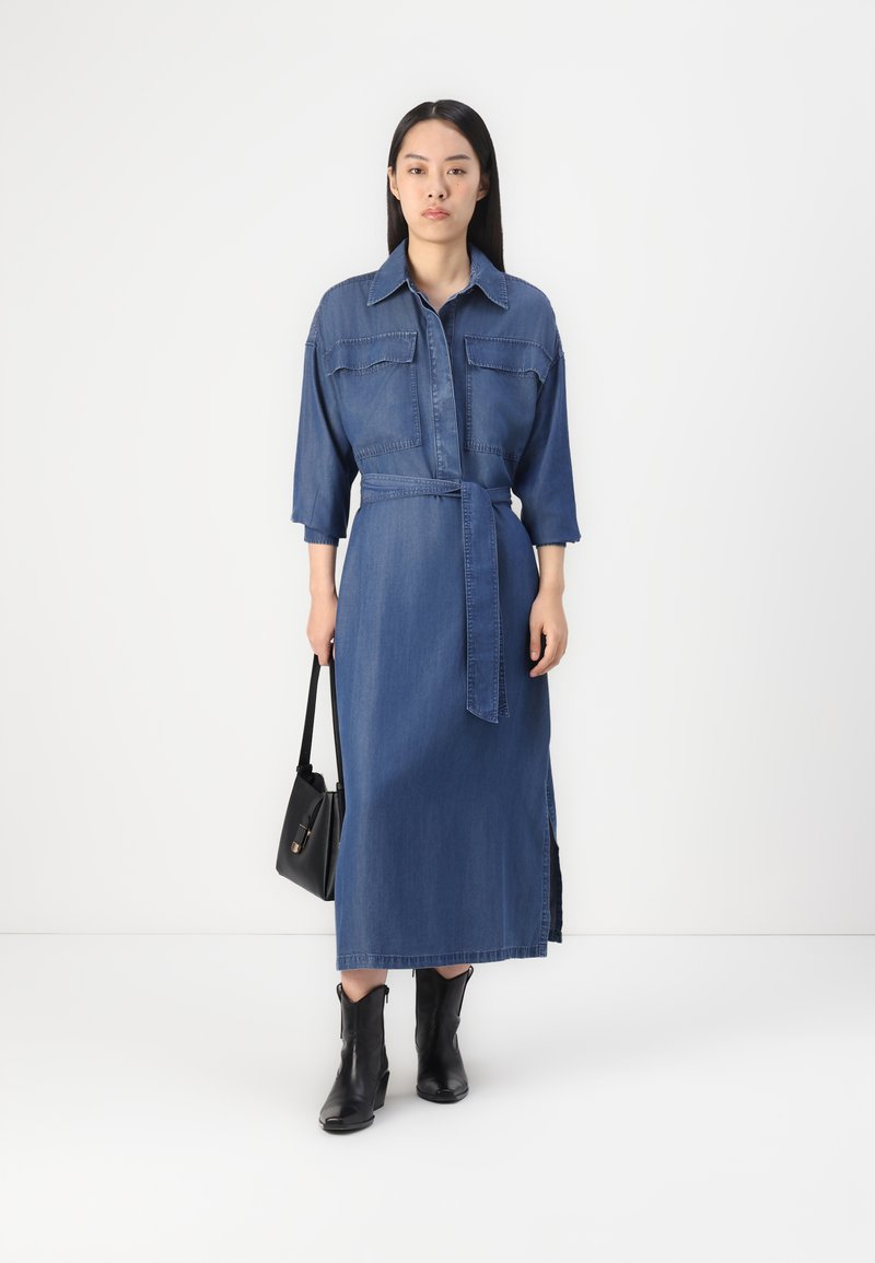 Robe chemise en denim bleu foncé avec une taille ceinturée, un col, deux poches poitrine et des fentes sur les côtés. Associée à des bottines noires et un sac.