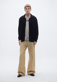 Tengerészkék kötött kardigán nagy gombokkal és zsebekkel, szürke póló fölött, bézs bő szárú nadrággal és barna cipővel párosítva.