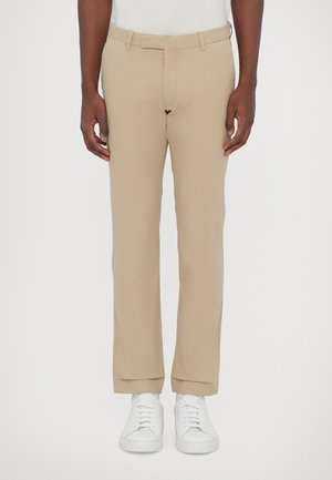 Man in beige broek en witte sneakers, staand tegen een effen lichte achtergrond, handen ontspannen langs het lichaam.