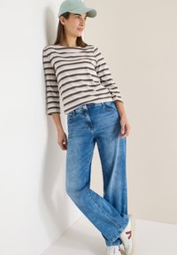 Femme portant un haut rayé à manches longues, un jean bleu, des baskets blanches et une casquette bleu clair, appuyée contre un mur blanc, regardant vers la droite.