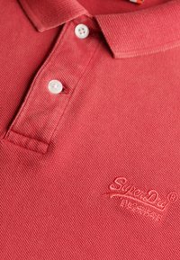 Polo rosso con tessuto a trama, caratterizzato da una chiusura con tre bottoni e un logo SuperDry ricamato nell'angolo in basso. Presenti bottoni bianchi.