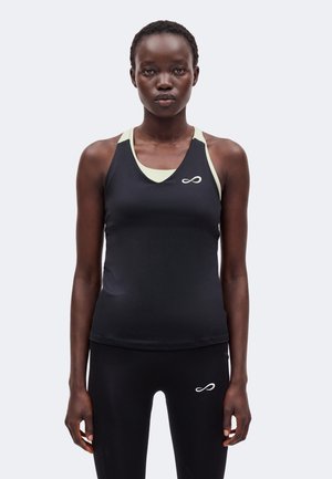 Mujer con camiseta deportiva negra sin mangas y leggings negros con símbolos de infinito blancos, de pie contra un fondo claro y uniforme.