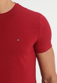 Nærbillede af en person iført en rød kortærmet t-shirt med rund hals og et lille Tommy Hilfiger-logo på brystet.