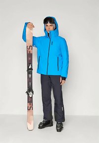 Jachetă de ski albastră cu fermoare negre, pantaloni de ski negri și bocanci de ski negri. Ținând schiuri bej cu legături. Ochelari gri pe cap.