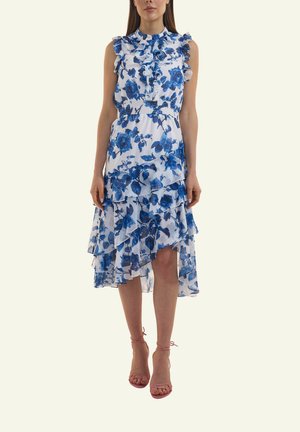 Frau trägt ärmelloses weißes Kleid mit blauem Blumenmuster, gestuftem High-Low-Saum und geraffter Taille, kombiniert mit rosa Riemchenabsätzen.