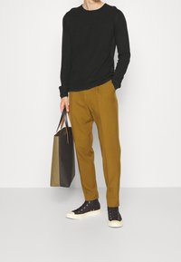 Homme portant une chemise noire à manches longues, un pantalon moutarde, des baskets montantes noires, tenant un sac fourre-tout bicolore avec une main dans la poche.