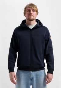 Gilet à zip navy avec une finition texturée, capuche ajustable et poches latérales, associé à un jean bleu clair. Design simple et fonctionnel.