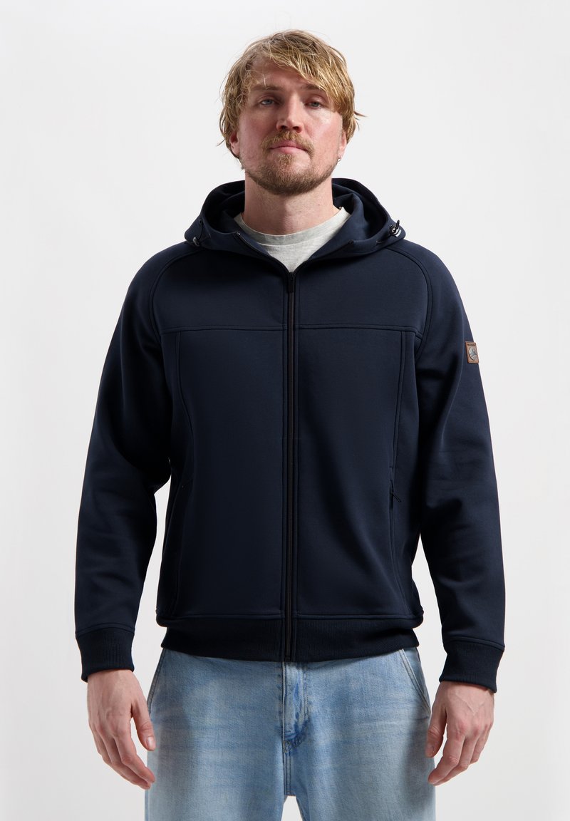 Gilet à zip navy avec une finition texturée, capuche ajustable et poches latérales, associé à un jean bleu clair. Design simple et fonctionnel.