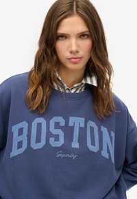Sudadera azul marino con "BOSTON" en letras bold azul claro, de corte relajado y mangas acampanadas, hecha de una tela suave.