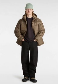 Braune Steppjacke mit Kapuze, getragen über einem schwarzen Hemd. Schwarze, locker sitzende Jeans und dunkle Schuhe. Hellgrüne, gerippte Mütze.