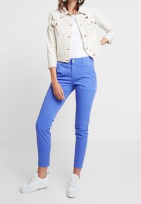 Giacca di denim chiaro sopra una maglietta bianca, abbinata a pantaloni blu aderenti e sneakers bianche. Presenta dettagli con bottoni e design a gamba stretta.