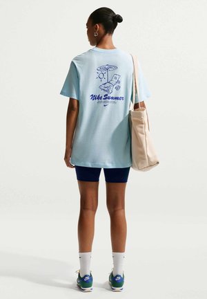 Γυναίκα που φοράει ανοιχτόμπλε μπλουζάκι Nike με καλοκαιρινό εκτύπωση παραλίας, σκούρα μπλε σορτς, άσπρες κάλτσες, πράσινα αθλητικά παπούτσια Nike και κρατάει μια μπεζ τσάντα ώμου.