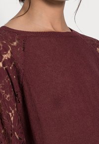 Bordeauxfarbener Pullover mit einem strukturierten Strickkörper und Spitzenverzierungen an den Ärmeln, die florale Muster und einen weiten Ausschnitt zeigen.