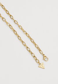 Gouden metalen ketting met grote ovale schakels, voorzien van een sluiting en een driehoekig label met het opschrift "GUESS." Gladde textuur en glanzende afwerking.