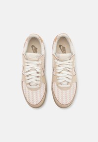 Une paire de baskets Nike beige et blanche avec une boîte à orteils à texture tissée, des lacets blancs et des accents beiges, vues d'en haut sur un fond blanc.