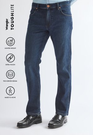 Homme portant un jean en denim bleu foncé et des chaussures en cuir noires, debout les mains dans le dos devant un fond clair uni.