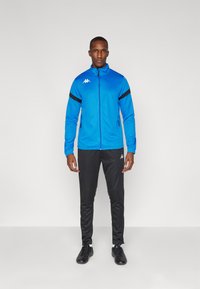 Veste zippée bleue avec des rayures noires sur les manches, associée à un pantalon noir. Présente le logo Kappa en blanc sur la veste et le pantalon.