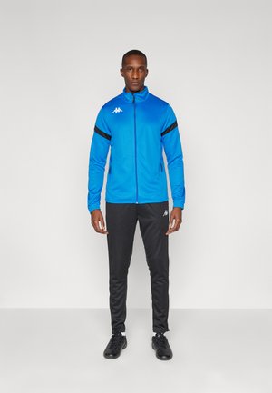 Blauwe zip-up jas met zwarte strepen op de mouwen, gecombineerd met zwarte broek. Bevat het Kappa-logo in het wit op zowel de jas als de broek.
