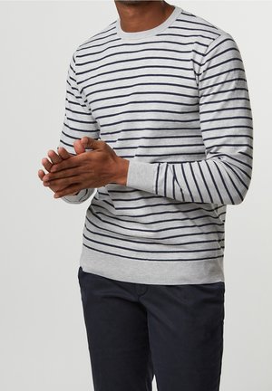 Man in gray long-sleeve shirt with black horizontal stripes and dark pants, clasping hands in front of torso against plain background.
