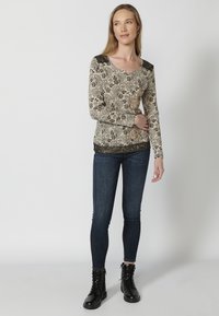Beige Langarmtop mit floralen Mustern; schwarze Spitzenakzente an den Schultern und am Saum. Kombiniert mit dunkelblauen Skinny-Jeans und schwarzen Schnürstiefeln.