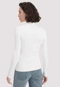 Weißes langärmliges Rippenoberteil mit hohem Kragen. Kombiniert mit blauen Jeans, die eine figurbetonte Form und glatte Textur zeigen.