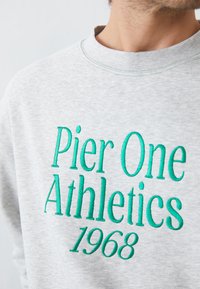 Szürke pulóver, amelyen boldog teal hímzés olvasható: "Pier One Athletics 1968". Anyaga puha, kerek nyakú és laza szabású.