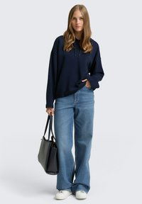 Felpa oversize blu navy con tasca a marsupio, abbinata a jeans a gamba larga di un blu chiaro e sneakers bianche, mentre tiene una borsa tote nera.