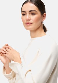 Witte blouse met een ontspannen pasvorm en geplooide mouwen. Model toont zilveren hoepeloorbellen en een neutraal make-up palet.