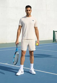 Mand i beige sportstøj med en tennisketcher og bold, stående på en blå tennisbane med net og hvid væg i baggrunden.