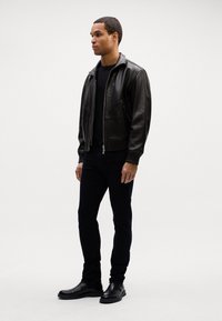Chaqueta bomber de cuero marrón con puños acanalados, camiseta de cuello redondo negra, jeans oscuros de corte ajustado y botas de cuero negras. Detalle de suela de goma visible.