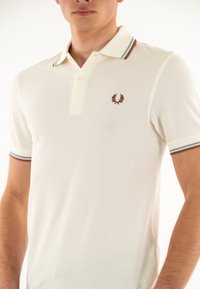 Creme polo skjorte i et struktureret stof. Den har en to-knaps placket, en kontraststribet krave og et broderet logo på brystet.