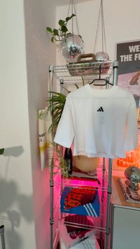 Valge lühendatud T-särk musta Adidas logo jaotisega, asetatud metallriiulile koos taime ja värviliste valgusaktsentidega.