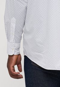 White shirt with small navy dots, long sleeves, and button cuffs. Fabric appears smooth, with a tailored fit and rounded hem. Worn with dark jeans.