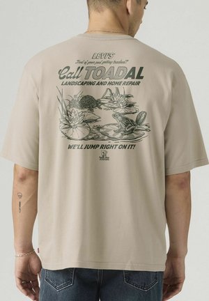 T-shirt beige avec impression verte présentant une tortue, une grenouille sur des nénuphars, et un texte promouvant des services d'aménagement paysager et de réparation domiciliaire.