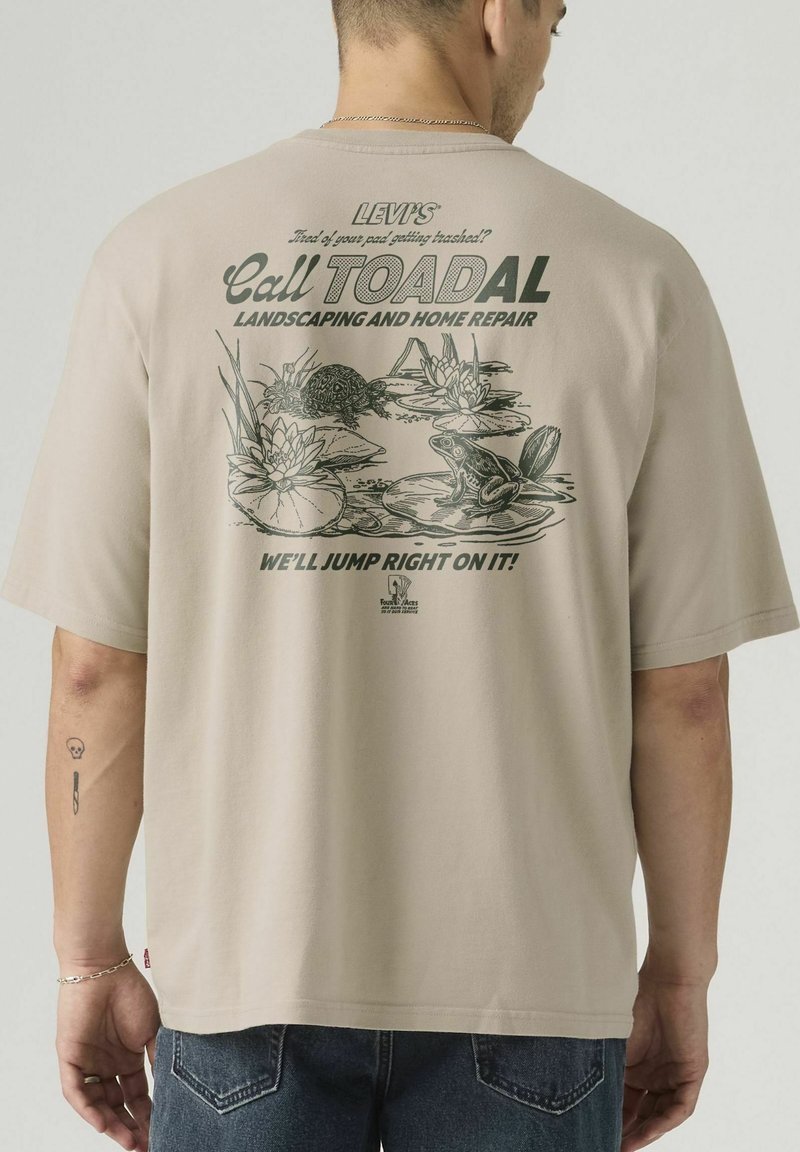 Maglietta beige con stampa verde raffigurante una tartaruga, una rana su foglie di ninfea, e testo che promuove servizi di giardinaggio e riparazioni domestiche.