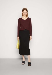 Top de manga longa canelado em burdeos com frente torcida, combinado com uma saia maxi de malha preta. Modelo segura uma pequena bolsa amarela. Sandálias pretas.